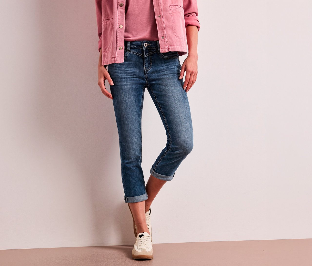 Une personne porte une veste rose, un haut rose, un Jean 7/8 Street One « Jane » bleu retroussé et des baskets claires.