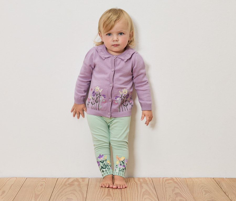 Une petite fille blonde se tient pieds nus devant un mur blanc, portant un cardigan lilas avec des fleurs et un legging vert avec des fleurs sur les jambes.