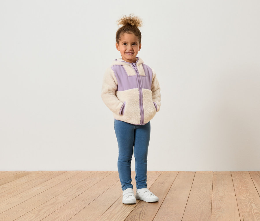 Une jeune fille se tient devant un mur blanc, vêtue d'une veste en polaire avec des accents violets, d'un jean bleu et de chaussures blanches.