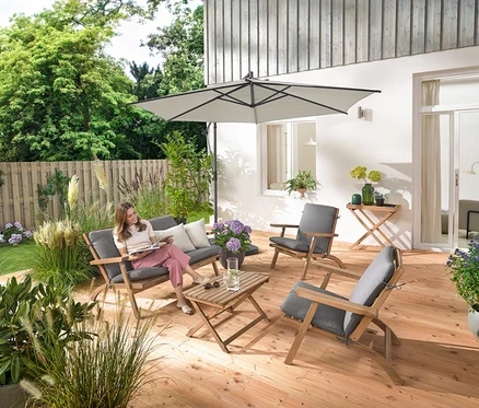 Une femme est assise sur un ensemble salon de jardin pliable avec coussins premium, elle lit et est entourée d'une tablette de jardin et de 2 coussins d’extérieur avec texture lin.