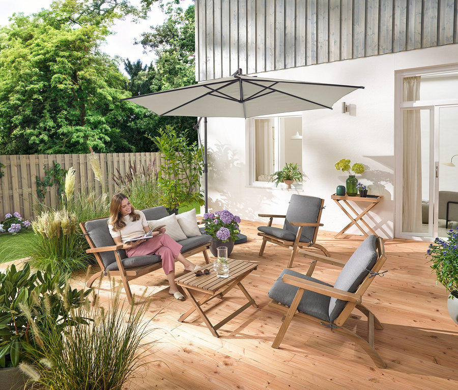 Une femme est assise sur un ensemble salon de jardin pliable avec coussins premium, elle lit et est entourée d'une tablette de jardin et de 2 coussins d’extérieur avec texture lin.