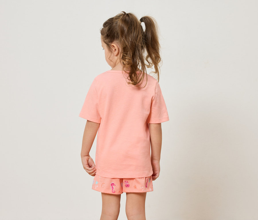 Vue de dos d'une petite fille avec des couettes portant un t-shirt orange et un short à motif d'hippocampes.