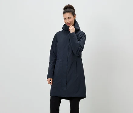 Une femme portant un chignon est vêtue d'un manteau bleu marine à capuche et d'un pantalon noir devant un fond neutre.