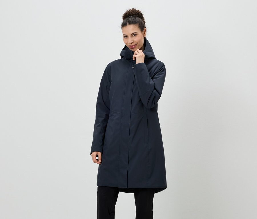 Une femme portant un chignon est vêtue d'un manteau bleu marine à capuche et d'un pantalon noir devant un fond neutre.
