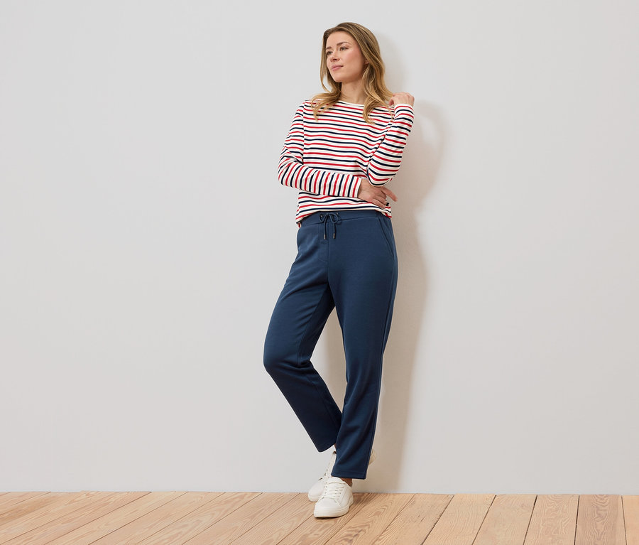 Une femme se tient devant un mur et porte un haut à manches longues rayé rouge et blanc et un pantalon de jogging bleu.