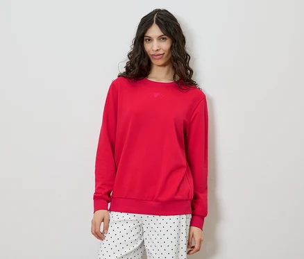 Une femme aux cheveux bruns bouclés porte un pull rouge et un pantalon à pois sur fond blanc.