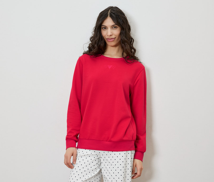 Une femme aux cheveux bruns bouclés porte un pull rouge et un pantalon à pois sur fond blanc.