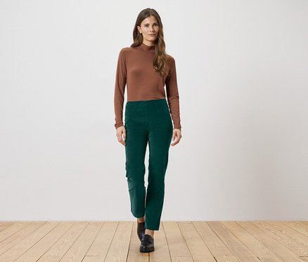 Une femme aux longs cheveux bruns porte un chemisier marron à manches longues et un pantalon vert. Elle se tient sur un plancher en bois.