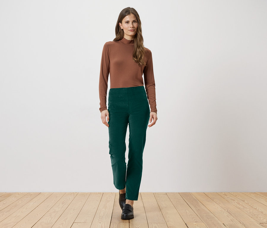 Une femme aux longs cheveux bruns porte un chemisier marron à manches longues et un pantalon vert. Elle se tient sur un plancher en bois.