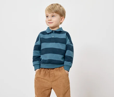 Un garçon blond se tient debout, les mains dans les poches, sur un fond blanc. Il porte un polo à rayures bleues et un pantalon en velours côtelé.