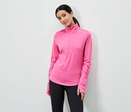 Une femme portant une queue de cheval porte un haut rose à manches longues avec fermeture éclair et un legging noir devant un mur blanc.