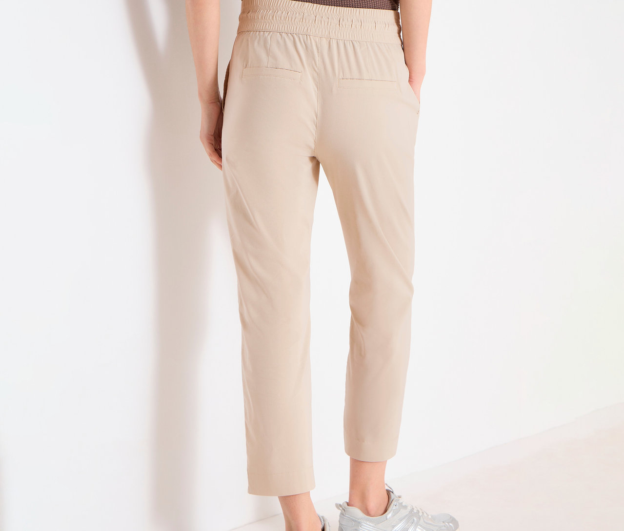Personne vue de dos portant un pantalon de jogging CECIL « Tracey » beige et des baskets argentées devant un mur blanc.