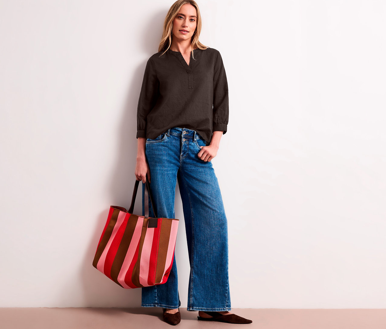 Une femme porte une blouse-tunique Street One gris foncé, un jean large et tient un sac cabas rayé rouge, rose et marron.