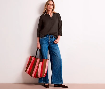 Une femme porte une blouse-tunique Street One gris foncé, un jean large et tient un sac cabas rayé rouge, rose et marron.