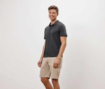 Un homme se tient debout et sourit, portant un polo anthracite et un short beige.