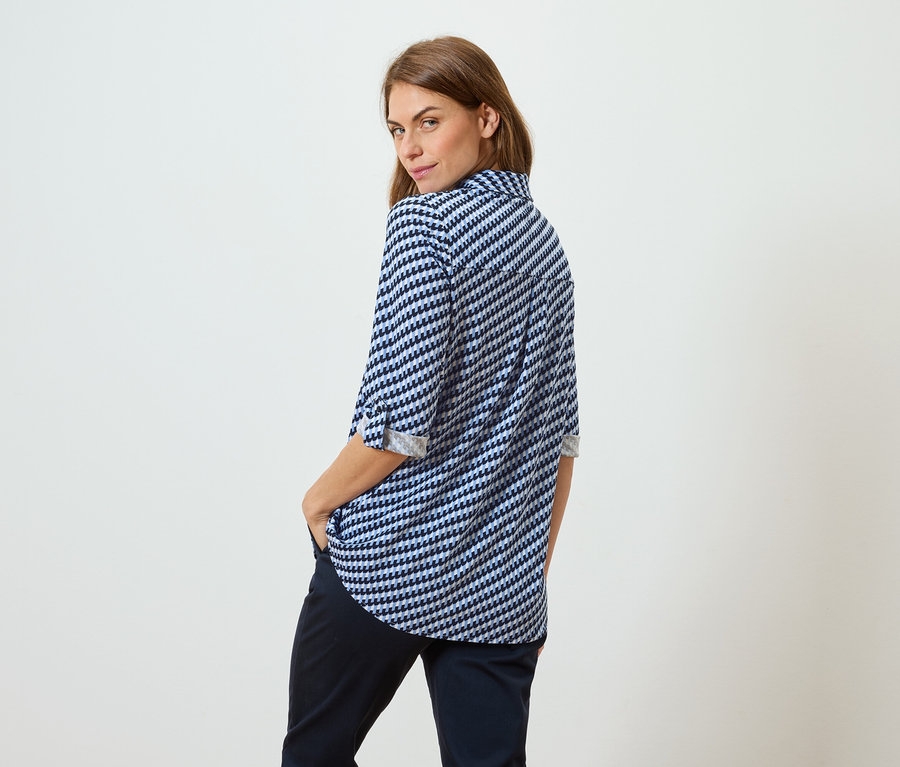 Une femme aux cheveux bruns portant un chemisier à motifs bleus et blancs et un pantalon foncé se tient dos au spectateur, regardant par-dessus son épaule.