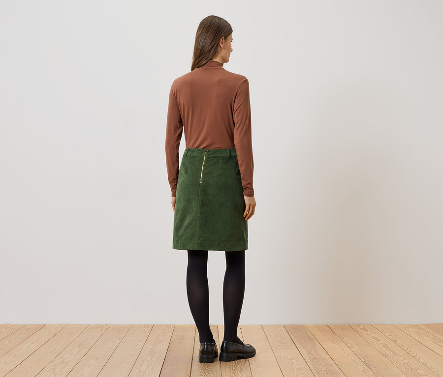 Une femme, vue de dos, porte un haut marron à manches longues, une jupe verte, des collants noirs et des chaussures noires.