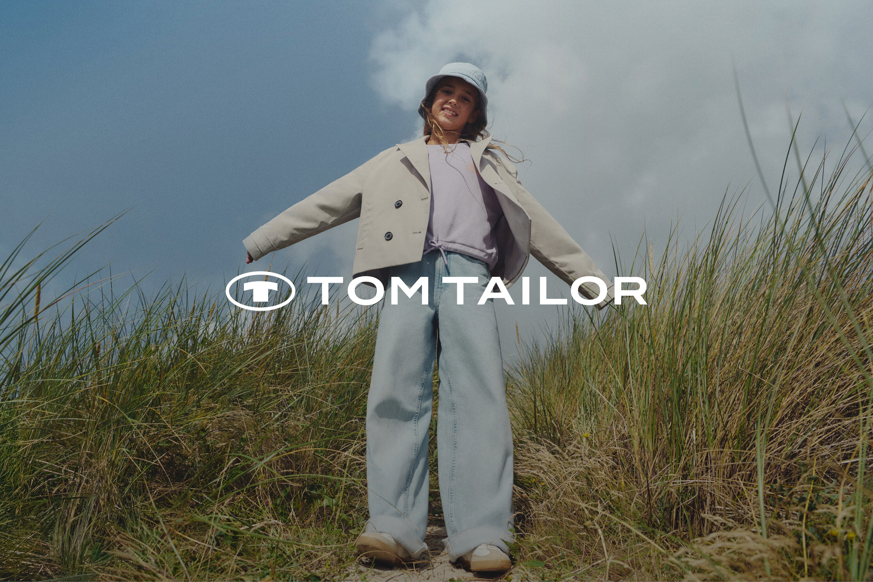 Une jeune fille se tient au milieu des herbes, portant une veste beige, un haut violet, un jean large et un chapeau bleu clair.