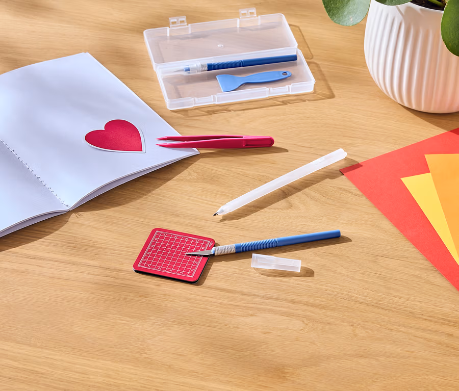 Sur une table en bois se trouvent un carnet ouvert avec un autocollant en forme de cœur rouge, une pince à épiler, un couteau de bricolage, un tapis de découpe et du papier coloré.