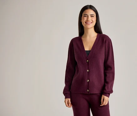 Une femme souriante aux longs cheveux foncés portant un cardigan bordeaux boutonné et un pantalon.