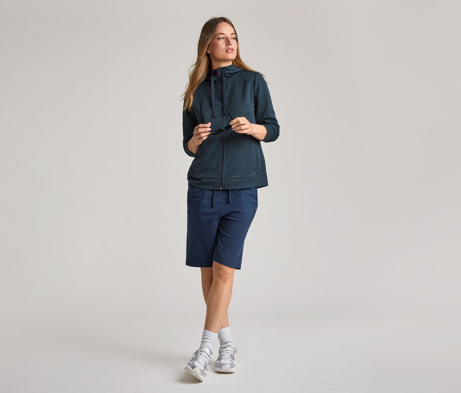 Femme portant une veste à capuche bleu marine et un short. Elle tient des lunettes de soleil.