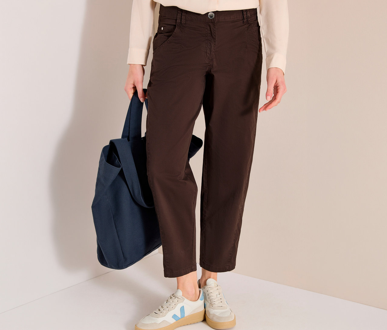 Une personne porte une chemise crème, un pantalon barrel marron CECIL « Scarlett » et des baskets claires avec des accents bleus, tenant un sac fourre-tout bleu foncé.