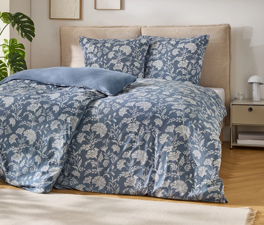 Un lit avec une parure de lit bleue à motifs floraux blancs et un oreiller bleu clair. Le lit a une tête de lit beige. À gauche se trouve une plante monstera, à droite une table de chevet avec une lampe et un verre. Un tapis beige est posé sur le parquet.