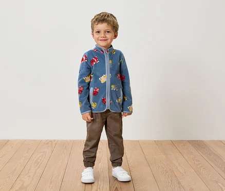 Un garçon se tient devant un mur, vêtu d'une veste polaire bleue à motifs de véhicules, d'un pantalon marron et de chaussures blanches.