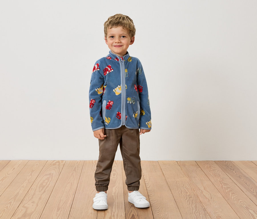 Un garçon se tient devant un mur, vêtu d'une veste polaire bleue à motifs de véhicules, d'un pantalon marron et de chaussures blanches.