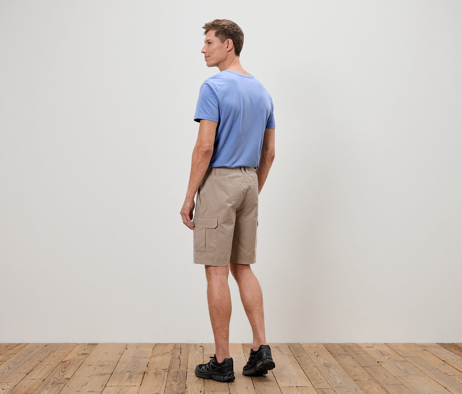 Un homme se tient dos à la caméra, il porte un t-shirt bleu, un short cargo beige et des baskets noires.