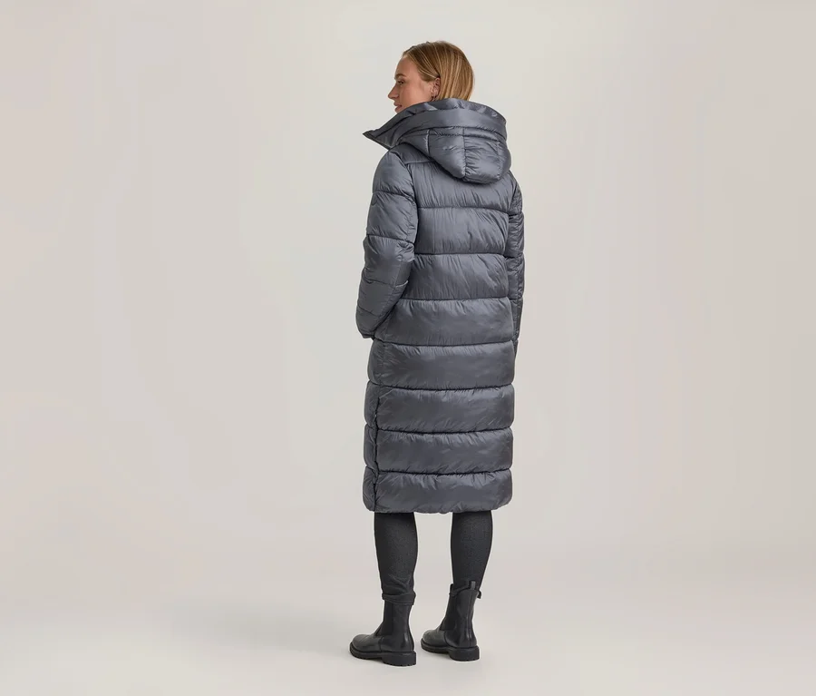 Femme vue de dos, légèrement tournée, portant un long manteau matelassé gris avec capuche, un pantalon foncé et des bottes noires.