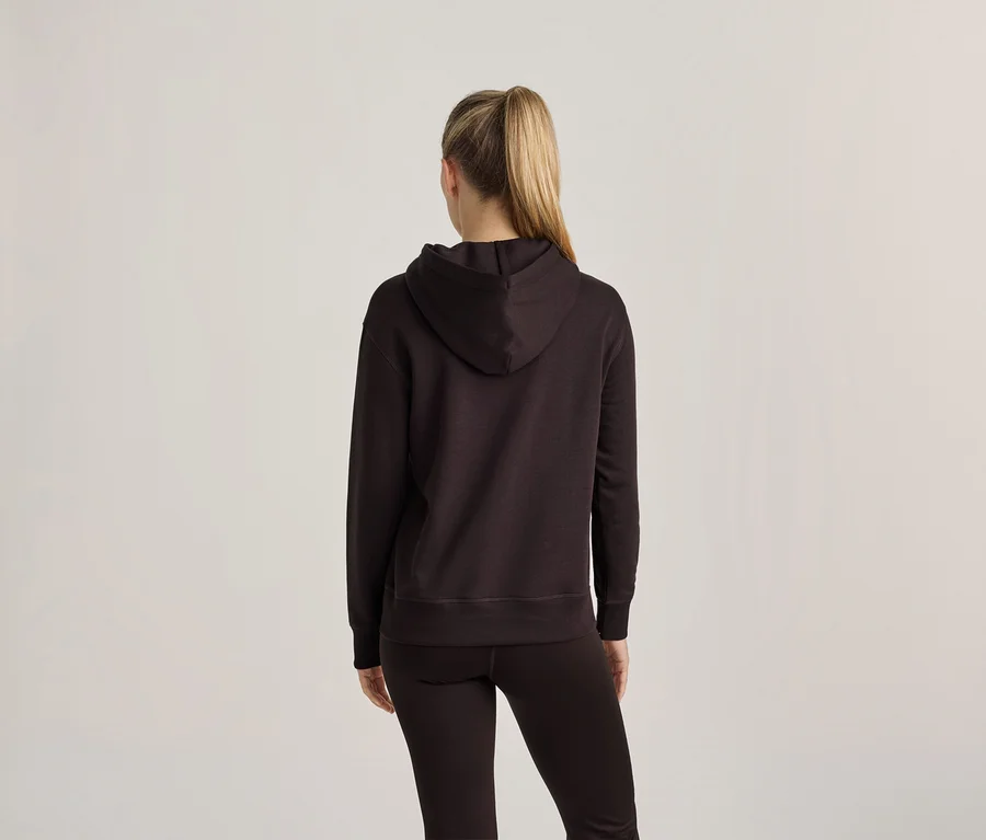 Femme vue de dos, portant un sweat à capuche foncé et des leggings foncés. Ses cheveux blonds sont attachés en queue de cheval haute.