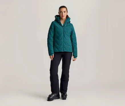 Femme debout portant une veste matelassée turquoise à capuche, un pantalon de ski noir et des chaussures de ski noires.