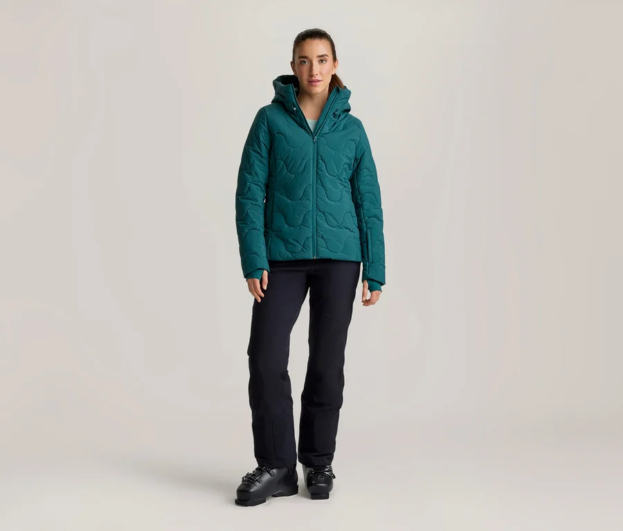 Femme debout portant une veste matelassée turquoise à capuche, un pantalon de ski noir et des chaussures de ski noires.