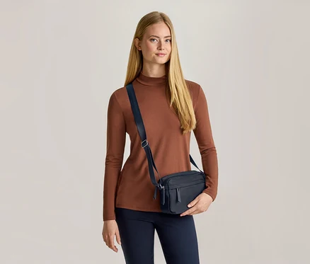 Une femme aux longs cheveux blonds porte un haut marron à manches longues, un pantalon bleu foncé et un sac bandoulière bleu foncé. Elle tient le sac de sa main gauche et regarde devant elle.