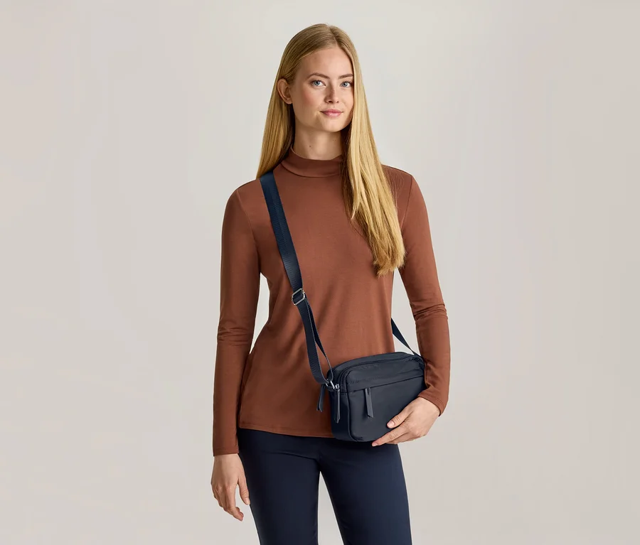 Une femme aux longs cheveux blonds porte un haut marron à manches longues, un pantalon bleu foncé et un sac bandoulière bleu foncé. Elle tient le sac de sa main gauche et regarde devant elle.