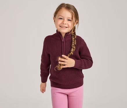 Jeune fille souriante avec une longue tresse, portant un pull côtelé bordeaux à demi-zip et un legging rose, tenant sa tresse de la main droite.