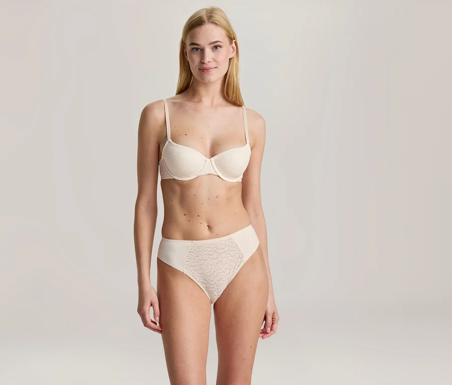 Femme blonde debout, souriante, portant un soutien-gorge beige clair et une culotte assortie avec des détails en dentelle.