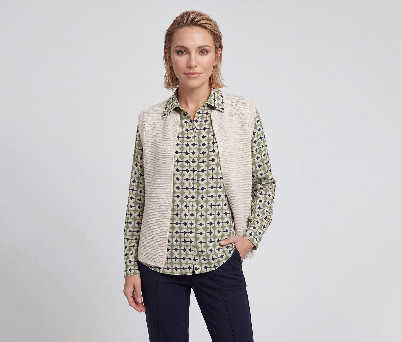 Femme aux cheveux blonds courts portant une chemise à motifs verts, beiges et bleu foncé, un gilet en maille beige et un pantalon bleu foncé, une main dans la poche.