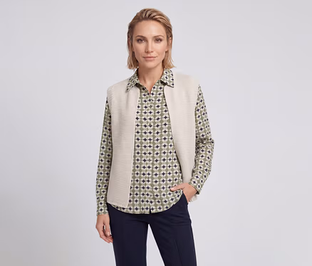 Femme aux cheveux blonds courts portant une chemise à motifs verts, beiges et bleu foncé, un gilet en maille beige et un pantalon bleu foncé, une main dans la poche.