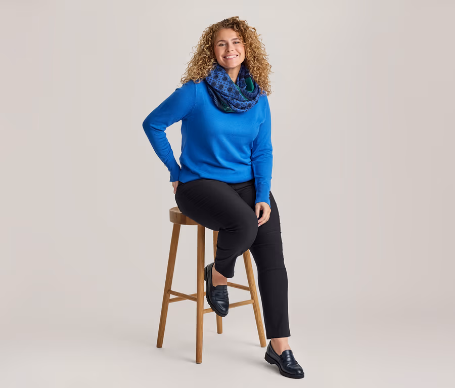 Femme souriante aux cheveux bouclés assise sur un tabouret en bois, portant un haut à manches longues bleu, un pantalon noir, des mocassins noirs et une écharpe tube à motifs bleus et verts.