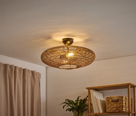 Un plafonnier ovale tressé éclaire une pièce, projetant des ombres sur le plafond. Un rideau marron clair est à gauche, et une étagère en bois avec des livres et un panier tressé est à droite.
