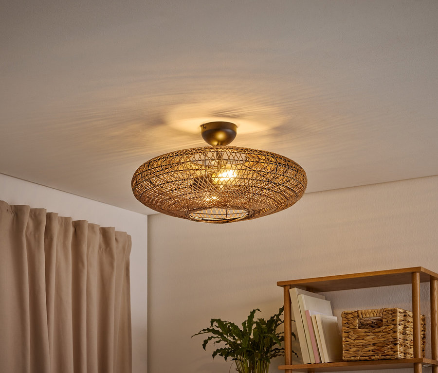 Un plafonnier ovale tressé éclaire une pièce, projetant des ombres sur le plafond. Un rideau marron clair est à gauche, et une étagère en bois avec des livres et un panier tressé est à droite.