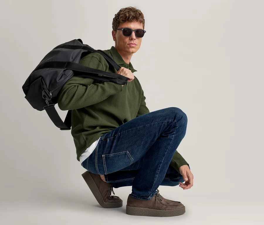 Homme accroupi, portant un polo vert foncé à manches longues, un jean bleu, des chaussures en daim marron et des lunettes de soleil, avec un sac de sport noir sur l'épaule gauche.