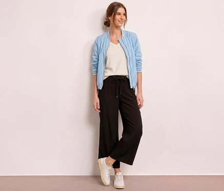 Une femme souriante se tient devant un mur clair, vêtue d'un blouson bomber bleu clair, d'un t-shirt crème et d'un culotte 7/8 Street One « EMMA » noire.