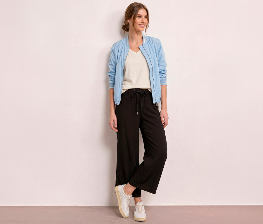 Une femme souriante se tient devant un mur clair, vêtue d'un blouson bomber bleu clair, d'un t-shirt crème et d'un culotte 7/8 Street One « EMMA » noire.