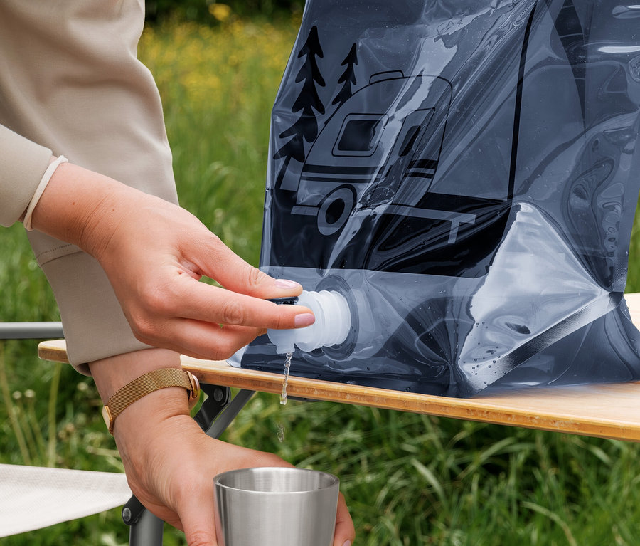 Une personne verse de l'eau d'un jerrycan pliable foncé et flexible avec un motif de camping-car dans une tasse en métal argenté. Le jerrycan est sur une table en bois en extérieur.