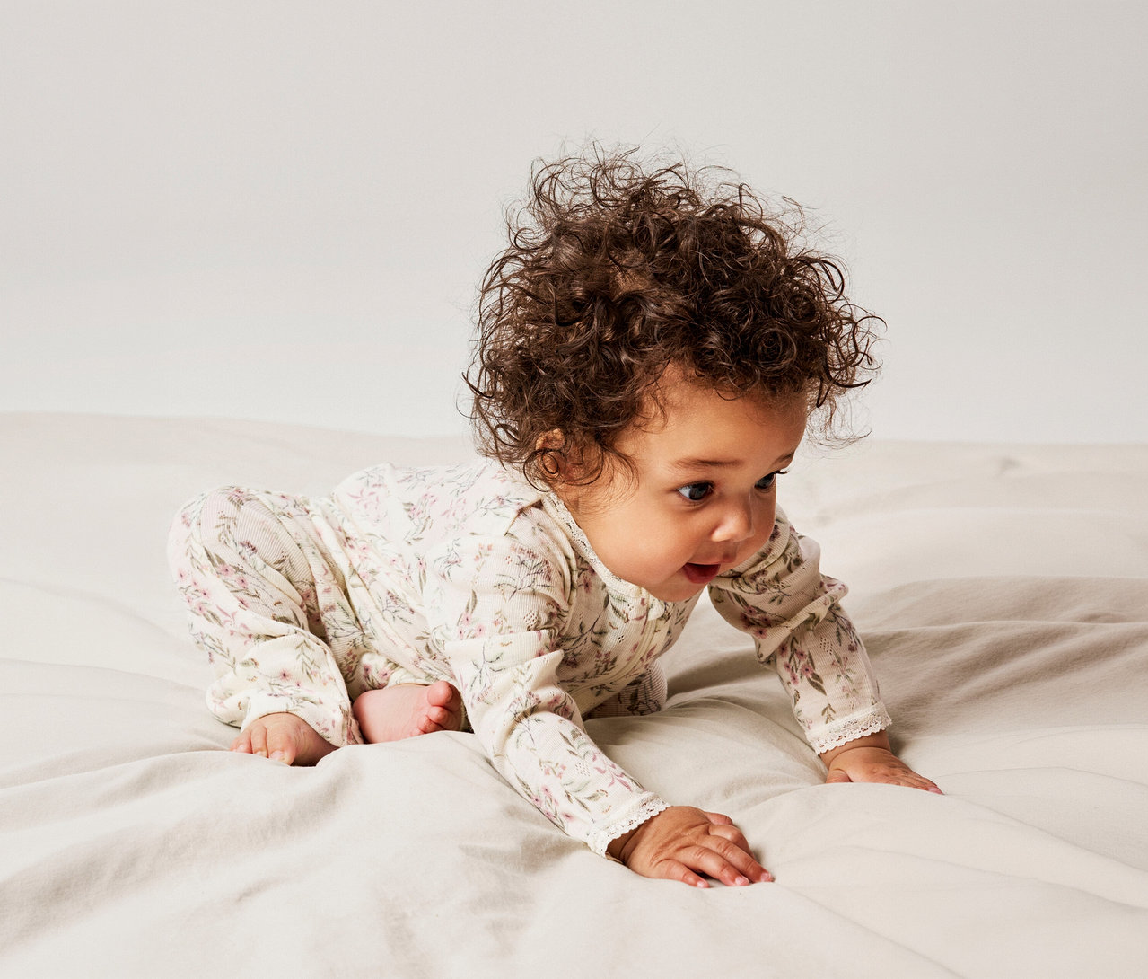 Un bébé aux cheveux bouclés foncés rampe sur un lit clair, vêtu d'une barboteuse florale crème, regardant vers l'avant avec la bouche ouverte.