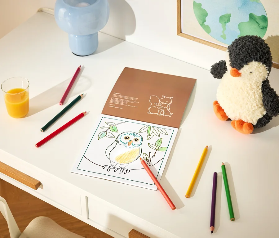 Un bureau blanc avec un livre de coloriage ouvert montrant un hibou sur une branche, partiellement colorié en jaune et bleu. Plusieurs crayons de couleur sont éparpillés autour du livre. À gauche se trouve un verre de jus d'orange, et à droite une peluche pingouin noire et blanche. Une lampe bleue et un tableau encadré de la Terre sont en arrière-plan.
