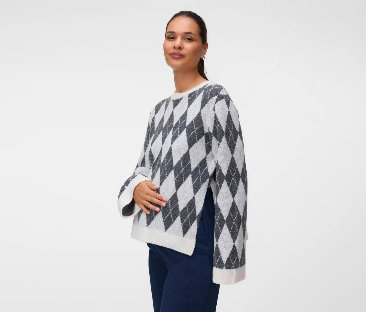 Femme portant un pull gris et blanc à motif losanges avec des fentes latérales et un pantalon bleu foncé, regardant la caméra avec une main sur son ventre.